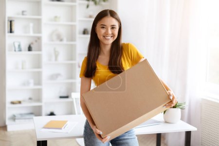 Téléchargez les photos : Heureuse femme asiatique tenant un grand colis en carton dans un bureau à domicile lumineux, coréenne souriant avec satisfaction alors qu'elle se prépare à expédier le paquet, montrant la livraison pour ses affaires en ligne - en image libre de droit