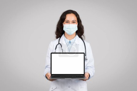 Téléchargez les photos : Médecin en masque de protection tenant un ordinateur portable avec écran blanc vierge, prêt pour les services de télésanté ou l'éducation des patients, fond gris, maquette - en image libre de droit