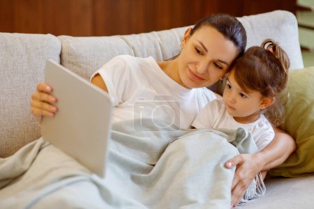 Photo for Caring mother and her little daughter with digital tablet relaxing on cozy couch, cuddled under a blanket mom and toddler child resting with modern gadget on comfortable couch at home, closeup - Royalty Free Image