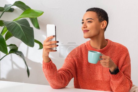 Téléchargez les photos : Femme souriante dans un pull orange profite d'un moment de loisirs, tenant une tasse de café dans une main et son smartphone dans l'autre, avec des plantes vertes en arrière-plan - en image libre de droit