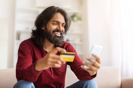 Téléchargez les photos : Homme indien positif profitant de la nouvelle application d'achat, faire une commande en ligne, assis sur le canapé à la maison, en utilisant un smartphone et tenant la carte de crédit, la banque virtuelle, payer sur Internet, e-commerce - en image libre de droit