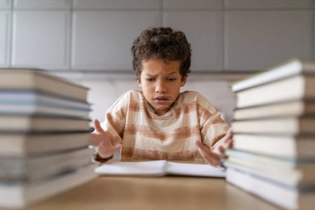 Photo for Puzzled black boy making homework surrounded by towering piles of books, stressed preteen male kid suffering confusion and puzzlement while looking in open workbook, having difficult task - Royalty Free Image