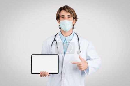 Photo for Male doctor in surgical mask presenting blank tablet screen, pointing finger at empty space for medical advertisement, recommending website, mockup - Royalty Free Image