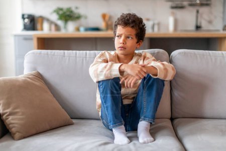 Photo for Pensive little boy with curly hair sitting on couch and looking away thoughtfully, preteen african american male child thinking about something, relaxing in living room interior, copy space - Royalty Free Image