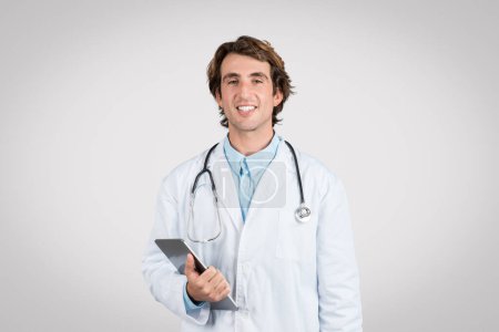 Téléchargez les photos : Médecin souriant portant un stéthoscope et un blouse de laboratoire tenant une tablette informatique, signifiant la disponibilité pour les services de soins de santé numériques, dans un contexte gris - en image libre de droit