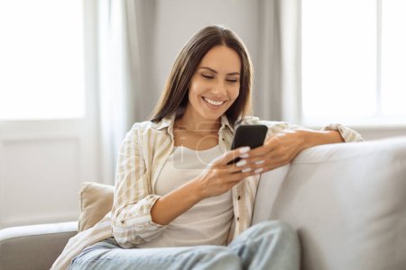 Téléchargez les photos : Jeune femme détendue souriante et naviguant sur son smartphone tout en s'allongeant sur le canapé, heureuse dame millénaire utilisant le téléphone mobile pour la communication en ligne ou les achats, reposant dans le salon - en image libre de droit