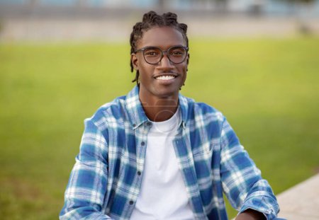 Téléchargez les photos : Joyeux jeune Afro-Américain avec des lunettes élégantes et chemise à carreaux bleus sourit à la caméra, assis à l'extérieur du campus universitaire. Portrait d'un étudiant masculin réussi à l'extérieur - en image libre de droit