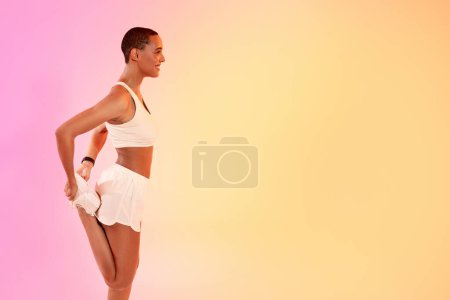 Foto de Una mujer latina sonriente con la cabeza afeitada estira la pierna detrás de ella, vistiendo un sujetador deportivo blanco y pantalones cortos, su perfil delineado contra un degradado de color rosa a amarillo de fondo - Imagen libre de derechos