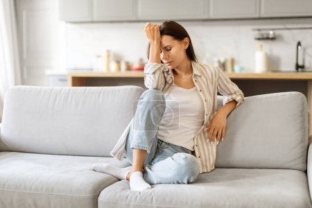 Photo for Depression Concept. Portrait Of Sad Young Woman Sitting On Couch At Home, Upset Millennial Female Feeling Lonely And Desperate, Touching Head With Hands, Crying In Living Room, Copy Space - Royalty Free Image