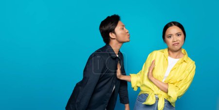 Asiatische Mann Millennial puckert um einen Kuss in Richtung einer unzufriedenen Frau, die den Vorstoß mit einer Handgeste ablehnt, sowohl vor einem expansiven türkisfarbenen Hintergrund, Studio