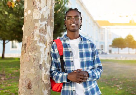 Téléchargez les photos : Un étudiant noir confiant appuyé contre un arbre posant avec un sac à dos et des lunettes, souriant à la caméra, exsudant une ambition éducative en dehors du campus universitaire le jour ensoleillé - en image libre de droit