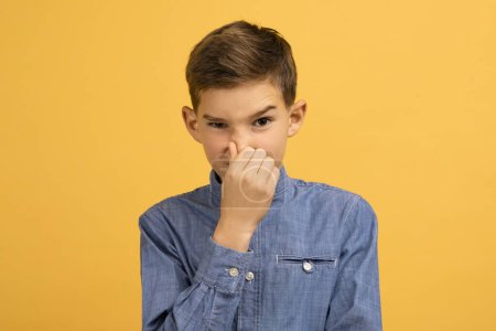 Displeased teen boy covering his nose to avoid bad smell, irritated teenage male child illustrating dislike or discomfort, standing isolated on yellow studio background, copy space