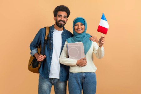 Téléchargez les photos : Éducation internationale. Couple étudiant arabe avec sacs à dos tenant le drapeau de France, Portrait d'un homme et d'une femme musulmans souriants dans le hijab avec des classeurs posant sur fond de studio beige, Espace de copie - en image libre de droit