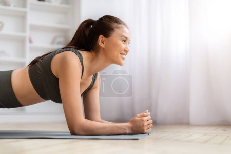 Foto de Concepto de entrenamiento en casa. Mujer asiática sonriente haciendo tablón de codo en estera de yoga en la sala de estar, mujer coreana deportiva haciendo rutina de ejercicios de fitness, ejercitándose en interiores, fortaleciendo sus músculos centrales - Imagen libre de derechos