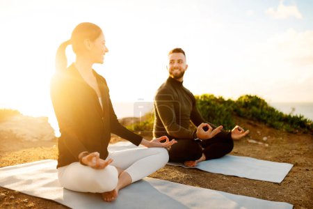 Téléchargez les photos : Homme et femme souriant, assis en position lotus sur des tapis de yoga, méditant à la lumière sereine du coucher de soleil sur une falaise surplombant l'océan - en image libre de droit