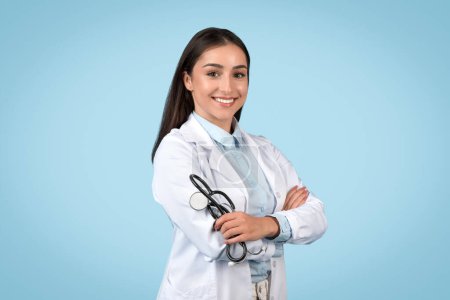 Joyeux jeune médecin caucasien porte un blouse et un stéthoscope, debout avec confiance sur un fond bleu apaisant