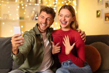 Téléchargez les photos : Jeune femme excitée et homme souriant prenant selfie comme elle montre sa main après la demande en mariage, avec des lumières festives en arrière-plan - en image libre de droit