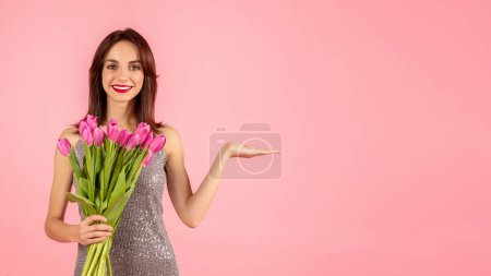 Photo for Smiling woman in a sparkling dress presenting with an open hand gesture while holding a bouquet of pink tulips, against a seamless pink studio background - Royalty Free Image