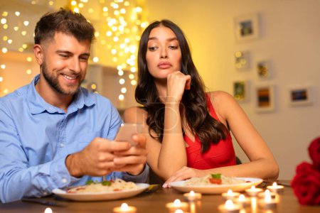 Engaged man browsing phone, disenchanted woman resting chin in hand, both at candlelit dinner, reflecting disconnect in modern relationships