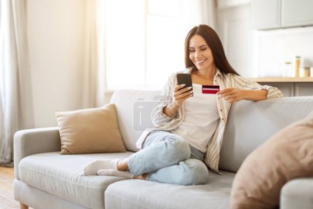 Photo for Relaxed young woman in casual clothes enjoying online shopping on her smartphone, smiling european female holding credit card, sitting on comfy sofa at home, enjoying mobile payments - Royalty Free Image