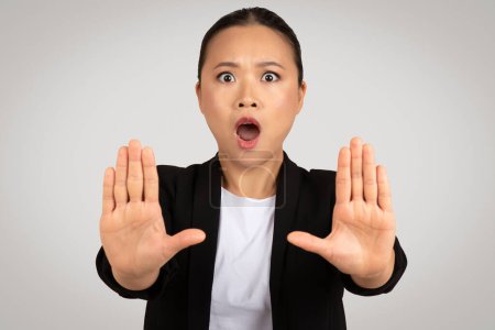 Foto de Sorprendido joven mujer asiática en traje con la boca abierta hacer gesto de parada con las manos, aislado sobre fondo gris, estudio. Di no, espacio personal, rechazo, lucha contra el acoso - Imagen libre de derechos
