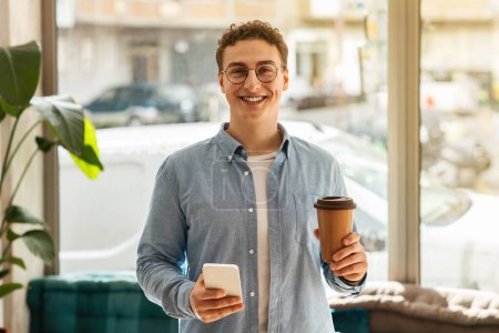 Photo for Young happy handsome european man student in glasses with coffee cup and smartphone in modern cafe, enjoy spare time. Break, lifestyle with device, ad and offer, work and study - Royalty Free Image