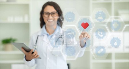 Foto de Tecnologías modernas en medicare, salud y gadgets. Mujer europea alegre en médico pelaje blanco con tableta digital en la mano tocando el botón rojo del corazón en la pantalla, panorama, espacio de copia - Imagen libre de derechos