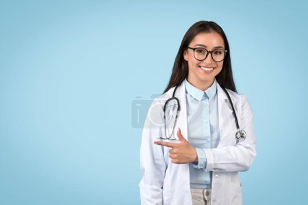 Lächelnde junge Ärztin in weißem Laborkittel macht eine wegweisende Geste, möglicherweise in Richtung ärztlicher Beratung oder Dienste auf blauem Hintergrund.