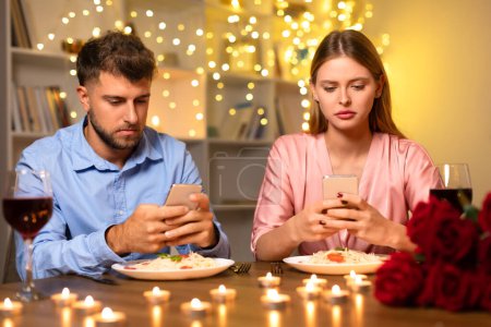 Man and woman engrossed in smartphones, ignoring each other over a romantic candlelit dinner with wine and pasta, intimacy ignored for tech
