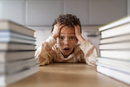 Photo for Stressed black boy surrounded by towering stacks of books, overwhelmed african american male kid touching head in shock and looking at camera, suffering stress and academic pressure, closeup - Royalty Free Image