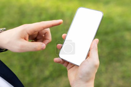 Foto de Primer plano de una persona manos sosteniendo teléfono inteligente con pantalla blanca en blanco, apuntando con el dedo, con fondo de hierba verde, cerca, recortado. Aplicación, sitio web, recomendación de blog - Imagen libre de derechos