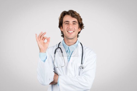 Selbstbewusster Arzt mit Stethoskop um den Hals, der vor grauem Hintergrund eine Okay-Geste macht, die auf eine erfolgreiche Behandlung oder Patientenzufriedenheit hinweist