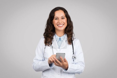 Téléchargez les photos : Médecine joyeuse en blouse de laboratoire utilisant un smartphone, gérant potentiellement des rendez-vous ou des communications avec les patients, sur un fond gris - en image libre de droit