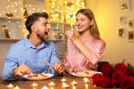 Photo for Cheerful woman feeding her partner pasta, both enjoying romantic dinner by candlelight with bouquet of roses and festive lights in the background - Royalty Free Image