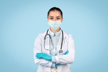 Téléchargez les photos : Médecin féminin avec une expression sérieuse, portant un masque chirurgical et des gants pour la sécurité sanitaire, symbolisant les précautions contre la pandémie, fond bleu - en image libre de droit