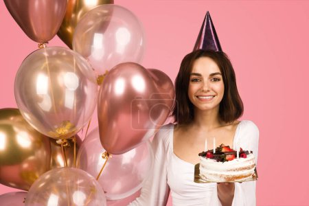 Photo for Radiant young caucasian woman in a white blouse and party hat, joyfully holding a birthday cake with candles, surrounded by a cluster of shiny balloons on a pink background - Royalty Free Image