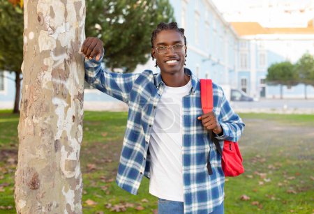 Téléchargez les photos : Étudiant afro-américain motivé en lunettes debout avec sac à dos par le bâtiment de l'université à l'extérieur, posant avec un sourire joyeux, incarnant l'esprit académique moderne - en image libre de droit