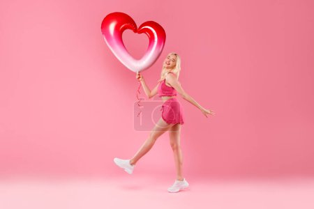 Foto de Chica rubia bailando alegremente, sosteniendo un globo de corazón rojo y blanco, en traje borroso rosa con zapatillas de deporte sobre un fondo rosa, sintiéndose eufórico, de cuerpo entero - Imagen libre de derechos