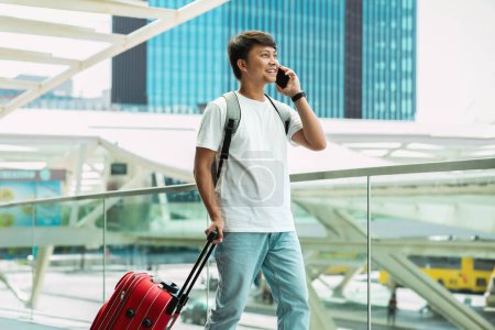 Photo for Roaming, stay connected while traveling. Asian guy tourist walking by airport, talking on phone, carrying red suitcase, copy space. Chinese young man traveler calling taxi - Royalty Free Image