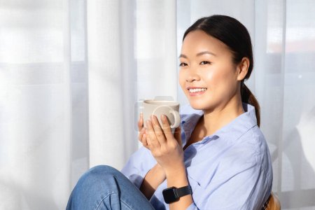 Foto de Primer plano de la mujer asiática bastante feliz millennial en ropa casual cómoda disfrutando de café aromático, sentarse en la silla al lado de la ventana en casa, sostener taza blanca, mirando el espacio de copia, sol bengala - Imagen libre de derechos