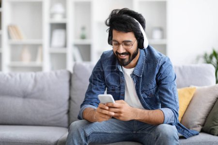 Photo for Relaxed indian man listening music on smartphone with wireless headphones, relaxing on couch at home, enjoying favorite songs on mobile phone or messaging with friends, resting on sofa in living room - Royalty Free Image