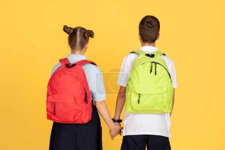 Téléchargez les photos : Deux enfants, une fille et un garçon, se tiennent le dos à la caméra, tenant la main et portant des sacs à dos aux couleurs vives, symbolisant l'amitié et un retour à l'école - en image libre de droit