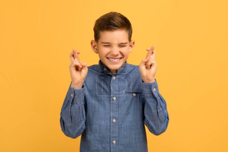 Cute Teen Boy Making Wish With Crossed Fingers And Closed Eyes, Superstitious Male Kid Hoping And Dreaming For Luck And Fortune, Standing Isolated On Yellow Studio Background, Copy Space