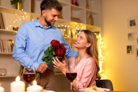 Photo for Smiling man presenting beautiful bouquet of red roses to surprised and happy woman during romantic candlelit dinner with wine, free space - Royalty Free Image