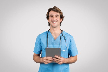 Cheerful male nurse in blue scrubs holding digital tablet, showcasing modern technology use in patient care and health management