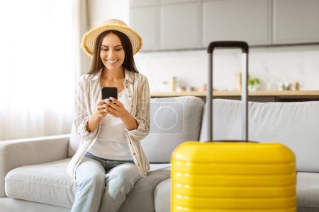 Téléchargez les photos : Femme souriante en chapeau de paille textant sur son téléphone, assise à côté de la valise jaune vif, heureuse femme signifiant joyeuse anticipation de voyage dans un cadre confortable à la maison, en utilisant l'application de réservation - en image libre de droit