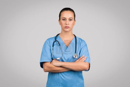 Téléchargez les photos : Infirmière déterminée en blouse bleue avec stéthoscope, bras croisés, faisant preuve d'une expression sérieuse de professionnalisme, sur fond gris - en image libre de droit