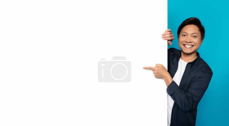 Photo for An Asian man with a cheerful smile is playfully peeking from behind a blank white vertical banner, pointing towards it, perfect for customizable content on a turquoise background - Royalty Free Image