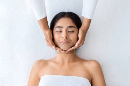 Photo for Top view masseuse making relaxing face massage for young pretty indian lady. Peaceful brunette woman getting lifting treatment at modern luxury spa in romantic atmosphere, white background - Royalty Free Image