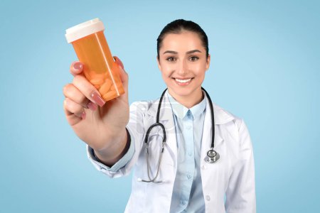 Foto de Médico sonriente que presenta frasco de pastillas, enfatizando la importancia de la adhesión de la medicación en el tratamiento, con estetoscopio, fondo azul - Imagen libre de derechos